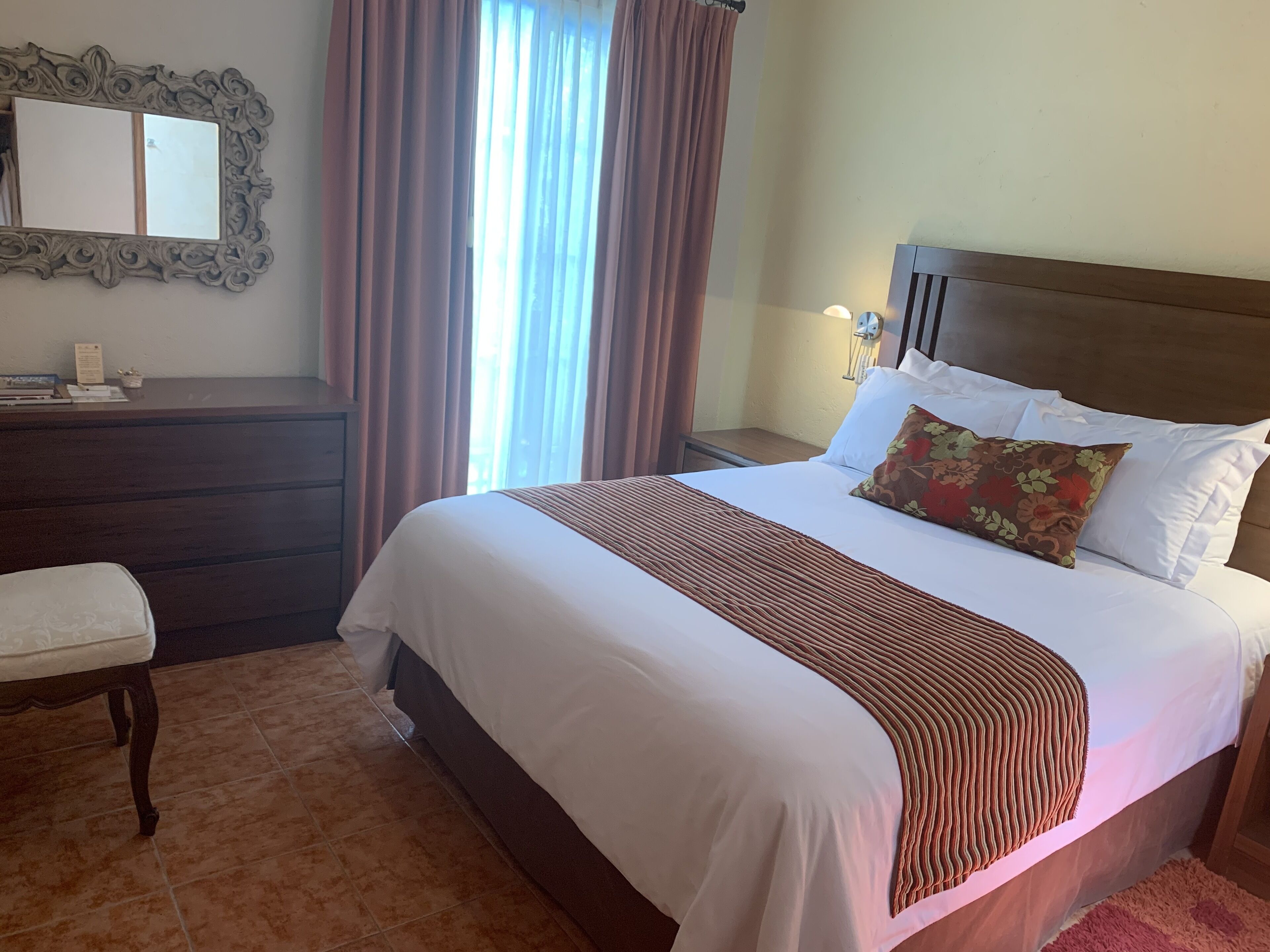 A hotel room with a large bed, a nightstand, a dresser, a mirror, and a window with curtains.