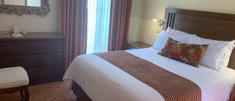 A hotel room with a large bed, a nightstand, a dresser, a mirror, and a window with curtains.
