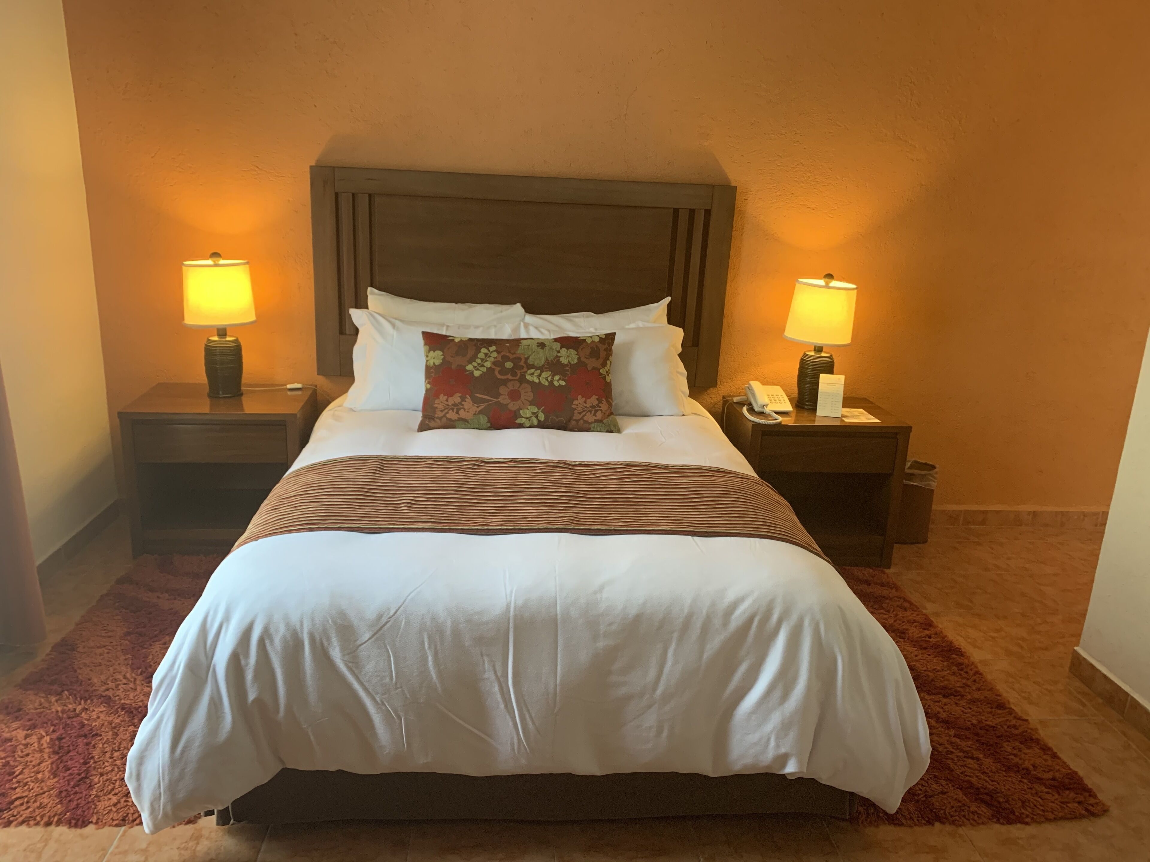 A neatly made bed with white linens, a striped throw blanket, and a floral pillow. There are two bedside tables with lamps, a phone, and a trash bin. The room has a warm, beige wall color and a brown carpet.