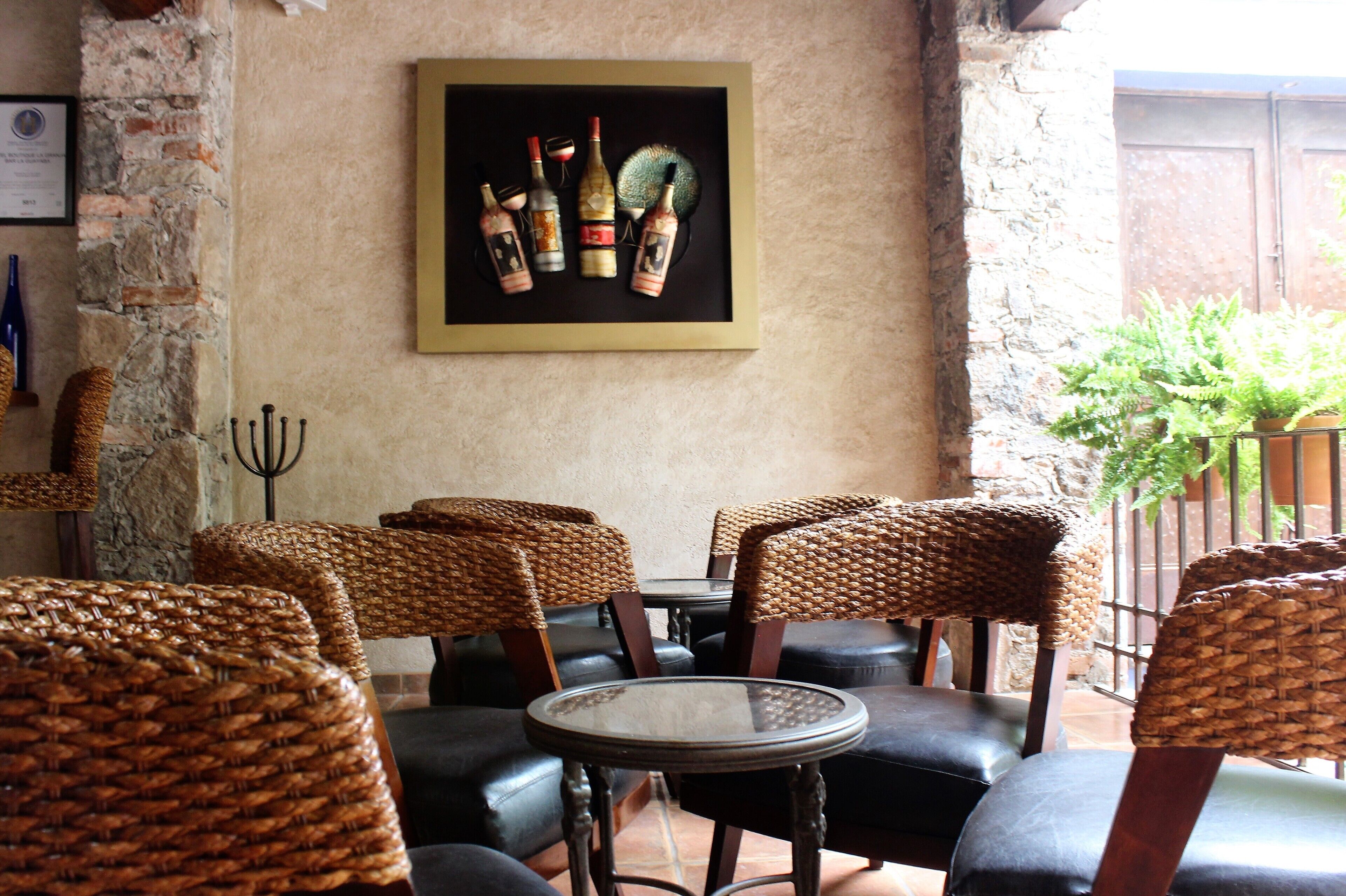 A restaurant interior with wicker chairs, a glass table, and a framed artwork of wine bottles on the wall.