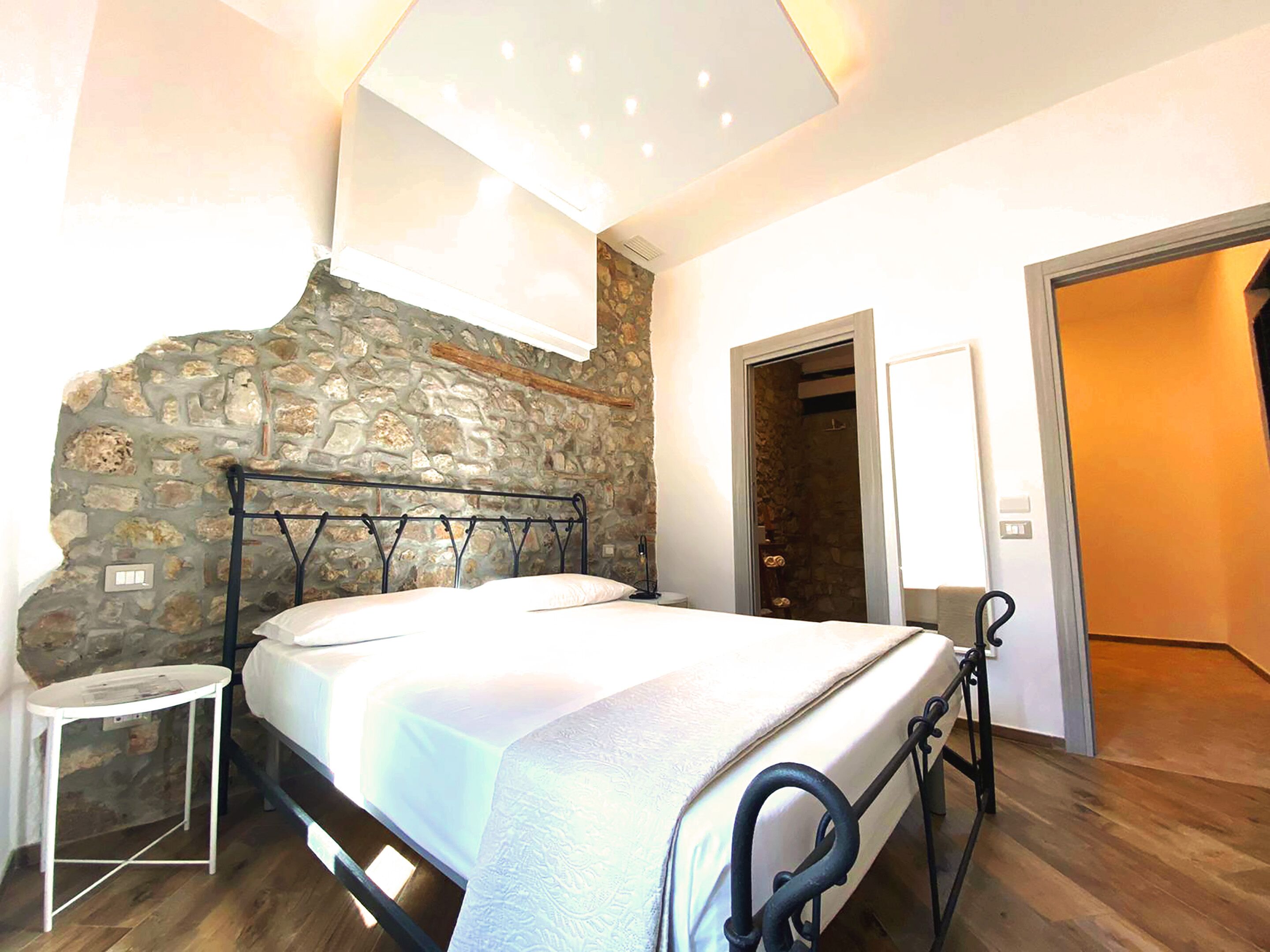 Un dormitorio con pared de piedra, cama con estructura metálica, mesita de noche pequeña y puerta que da a otra habitación.