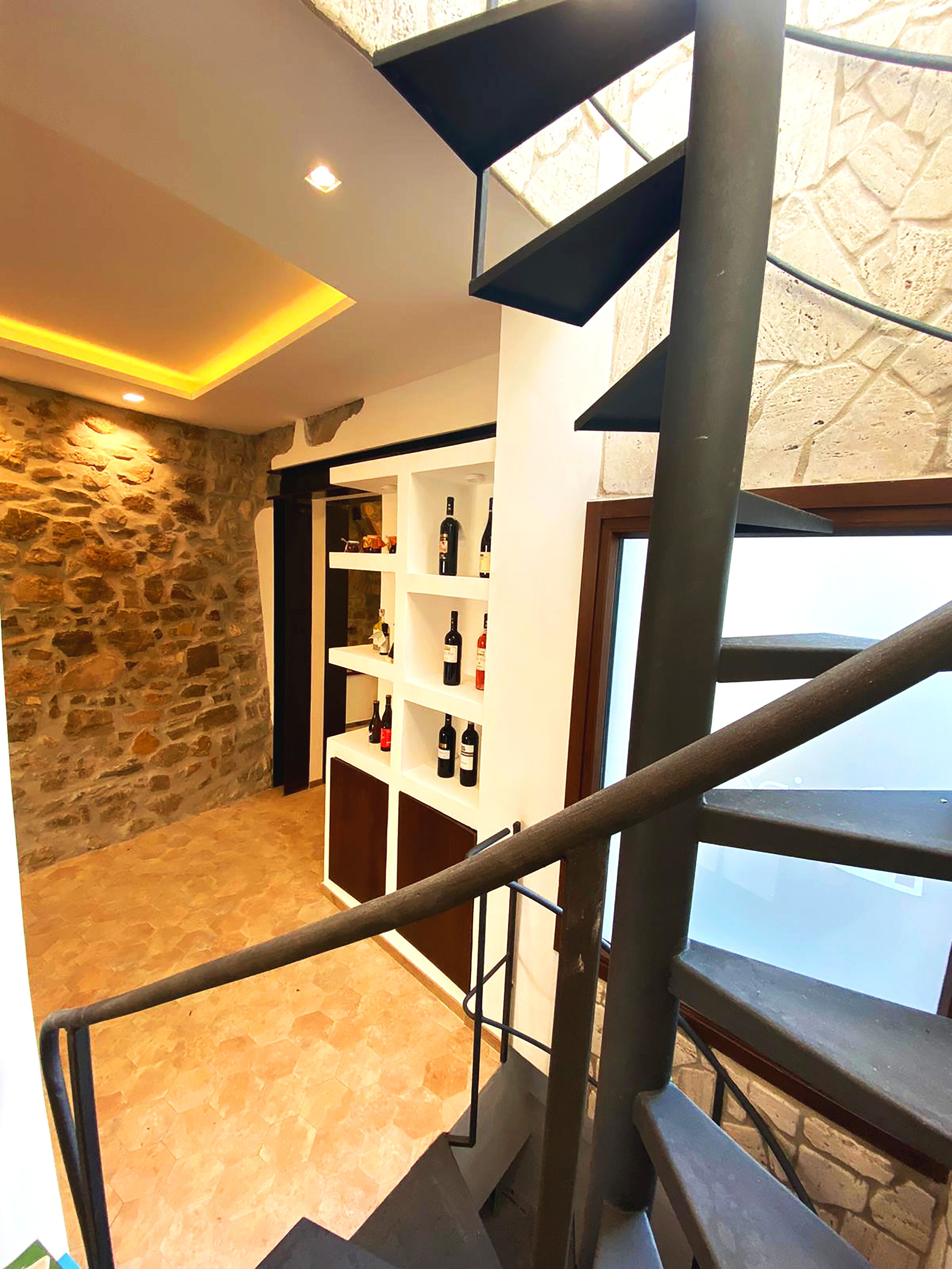 A modern interior with a stone wall, a wooden bookshelf with wine bottles, and a staircase with a metal railing.