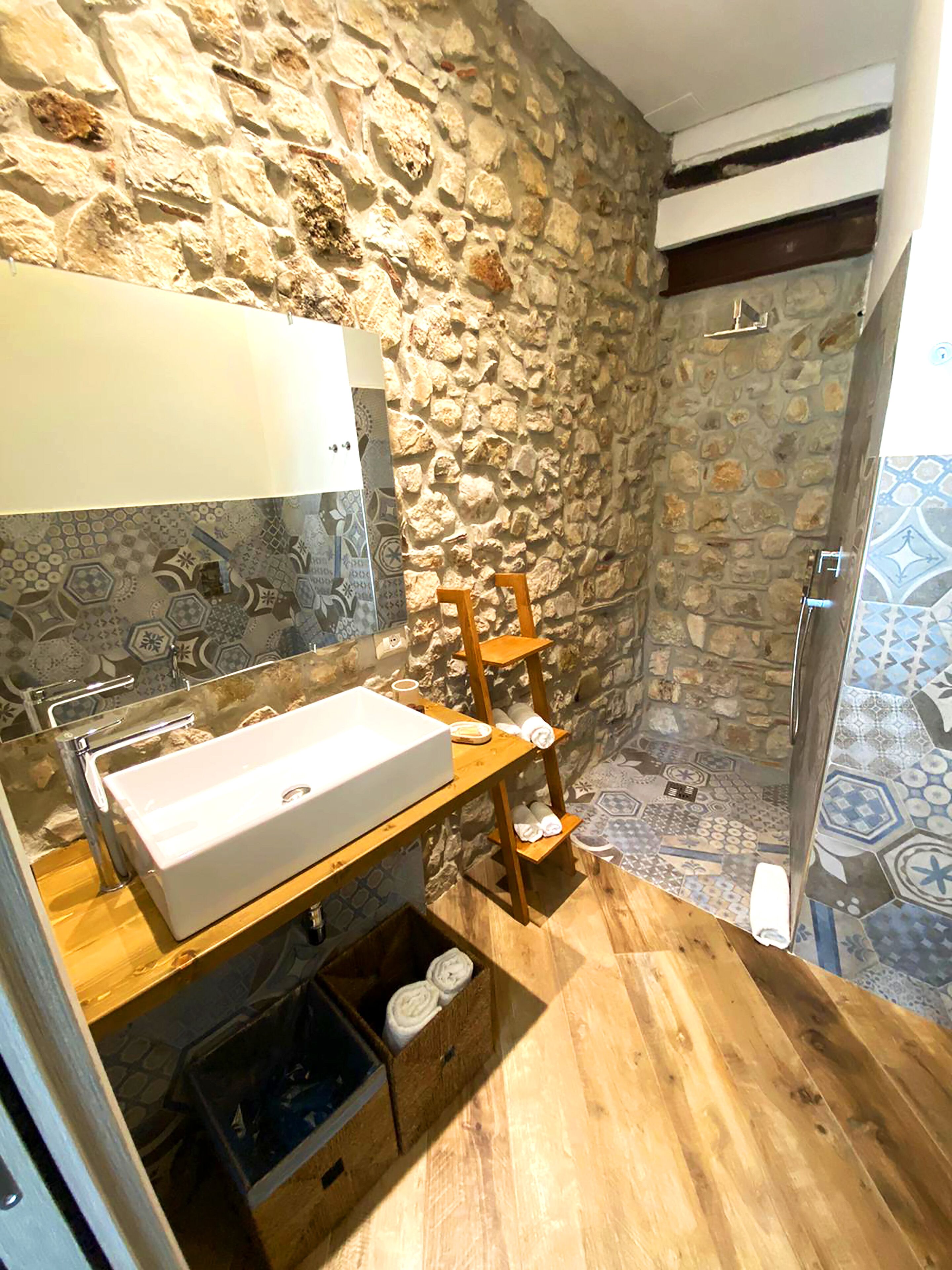 Un bany amb paret de pedra, una estanteria de fusta amb tovalloles, un lavabo blanc i una zona de dutxa.