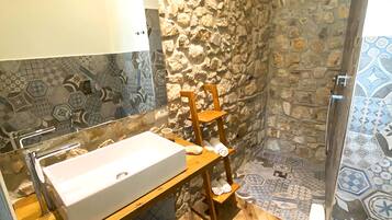 Un bany amb paret de pedra, una estanteria de fusta amb tovalloles, un lavabo blanc i una zona de dutxa.