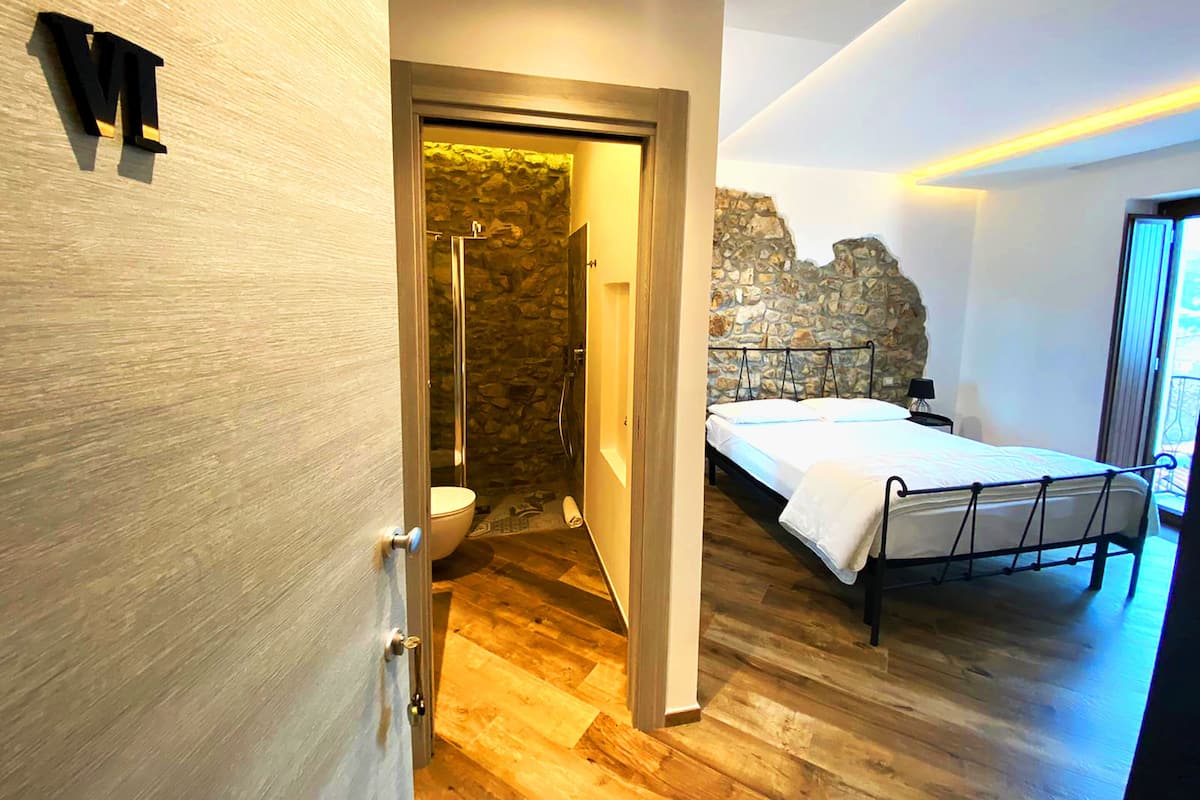 A modern bedroom with a wooden floor, a bed with a metal frame, a stone wall, and a bathroom with a stone shower enclosure.