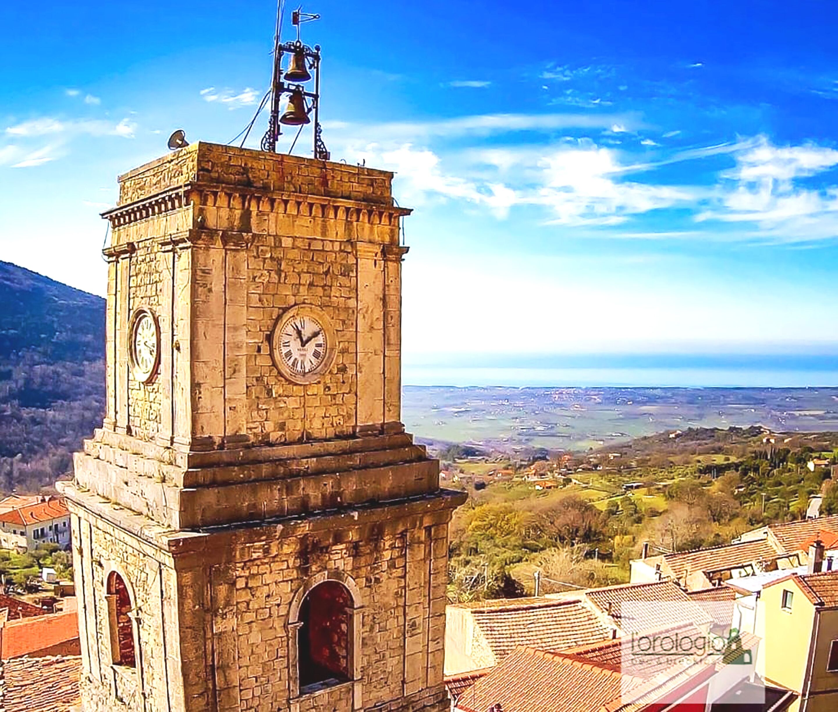 A stone clock tower with a weather vane, surrounded by a village with terracotta roofs.