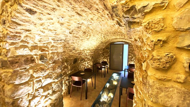 Interior de paredes de piedra con una larga mesa y sillas, una vitrina de vidrio y una puerta que da a otra habitación.