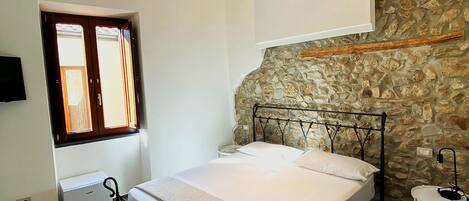 A bedroom with a stone wall, a bed with a metal frame, a small bedside table, and a window with wooden shutters.