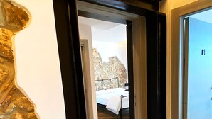 Un pasillo que conduce a un dormitorio con pared de piedra, suelo de madera y una cama con manta blanca.