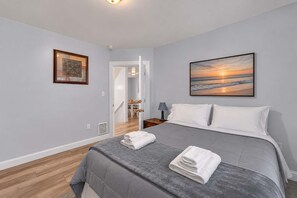 A bedroom with a bed, a painting of a sunset, a framed picture, and a view of a hallway. - Shortstop Apt 2 - All Star Baseball Rentals (Oneonta)
