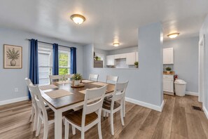 A dining area with a wooden table and white chairs, a kitchen with white cabinets, and a window with blue curtains. - Shortstop Apt 2 - All Star Baseball Rentals (Oneonta)