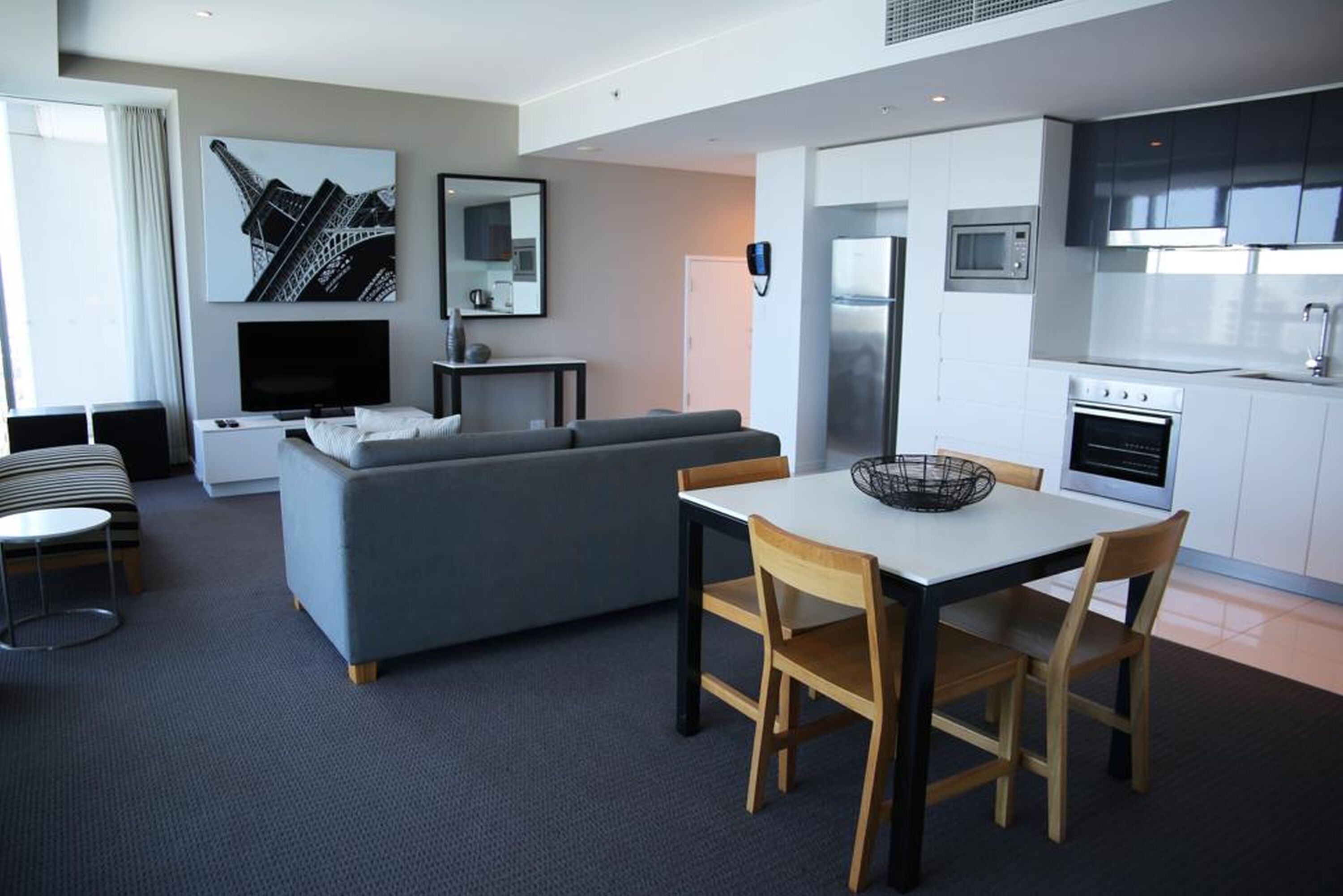 A modern hotel room with a grey sofa, a dining table with chairs, a flat-screen TV, a small side table, and a kitchen area with a refrigerator, microwave, and oven.