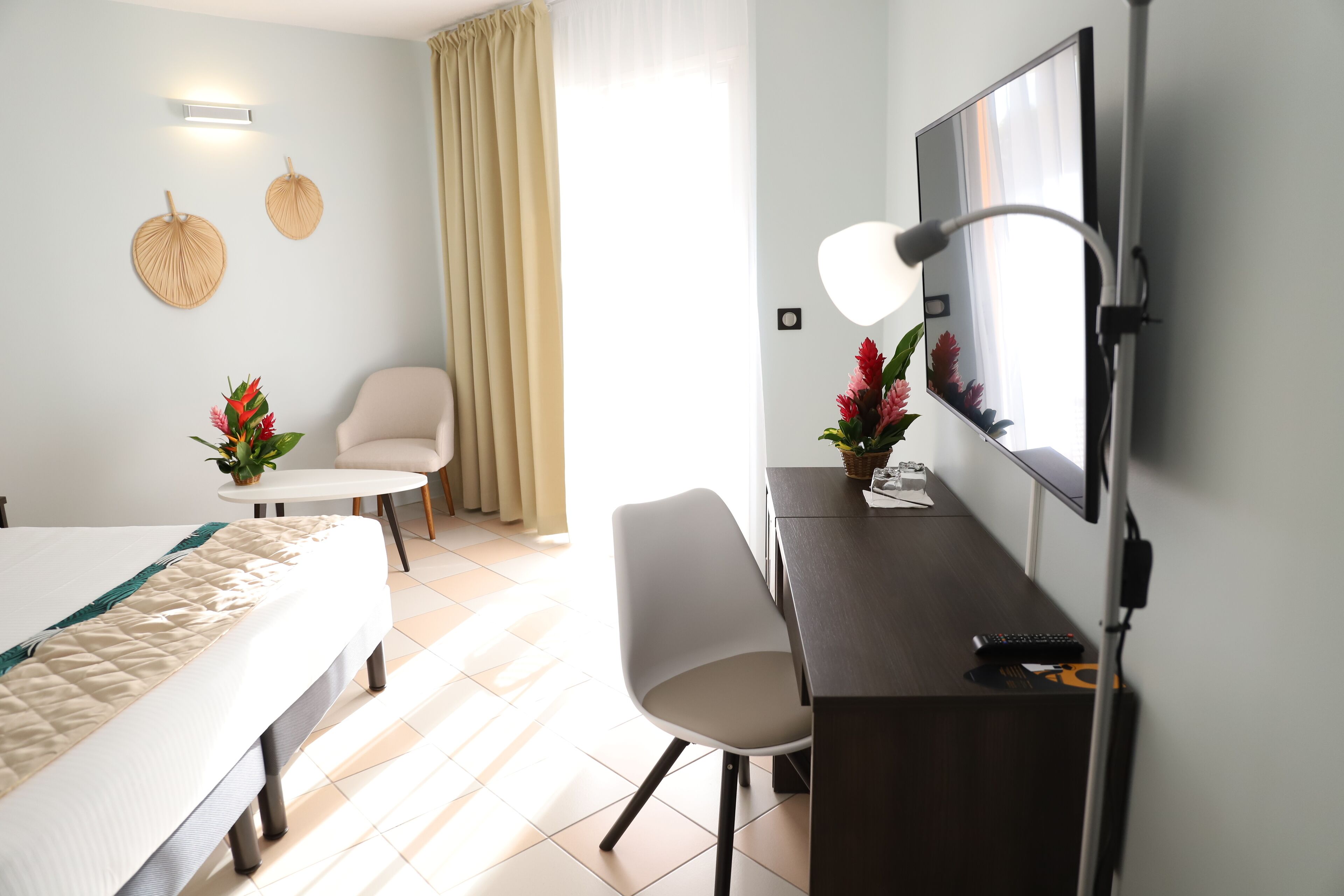 A hotel room with a bed, a desk with a computer and keyboard, a chair, a TV, and a window with curtains.