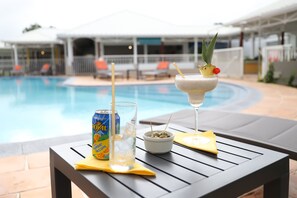 A poolside table with a can of Royal Hawaiian, a glass of iced drink, and a cocktail garnished with a pineapple and mint.