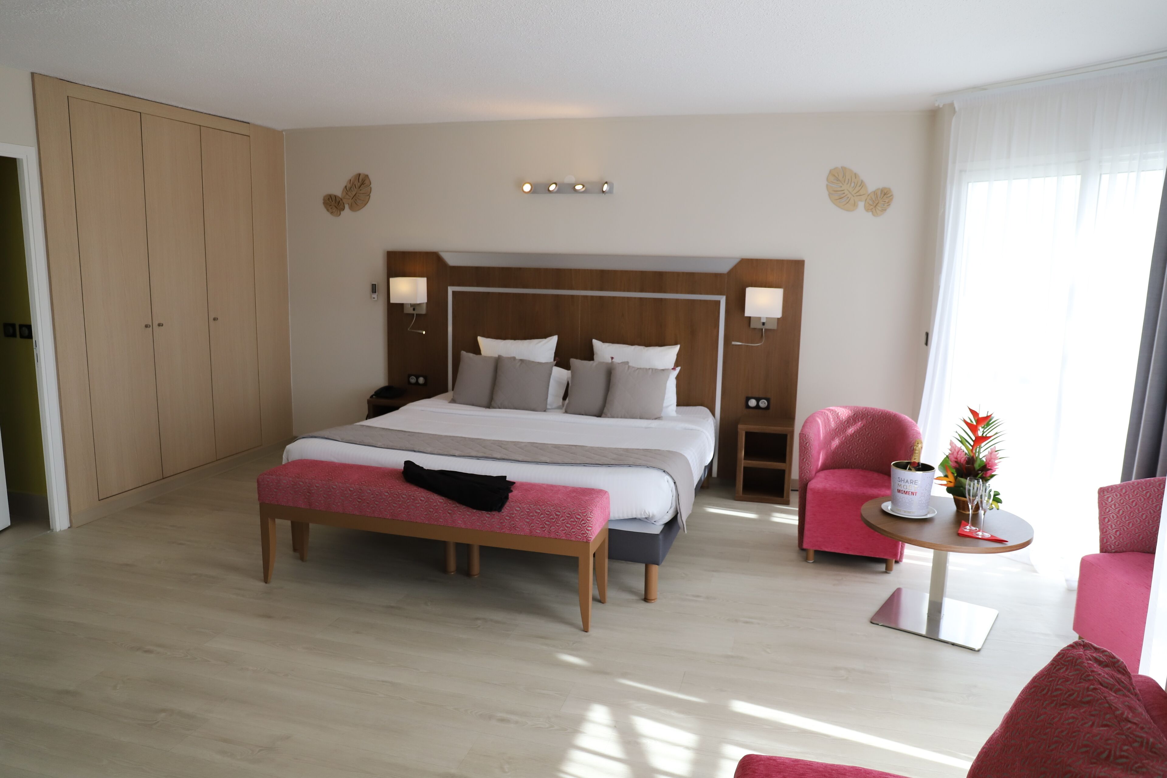 A spacious bedroom with a large bed, bedside lamps, a wooden wardrobe, a bench, and two pink armchairs.