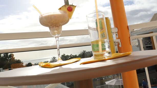 Un balcon avec une boisson et un verre d’eau, une piscine en arrière-plan et un ciel dégagé.