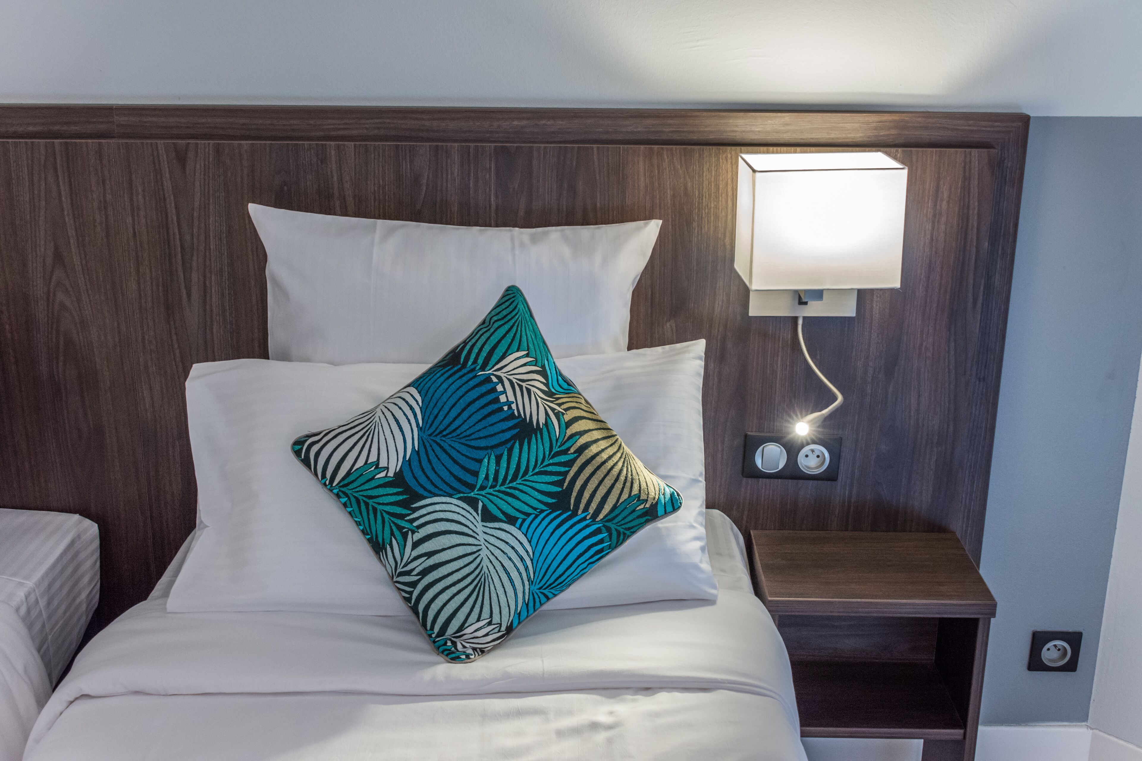 A neatly made bed with white linens, a teal leaf-patterned pillow, and a bedside table with a wall-mounted lamp.
