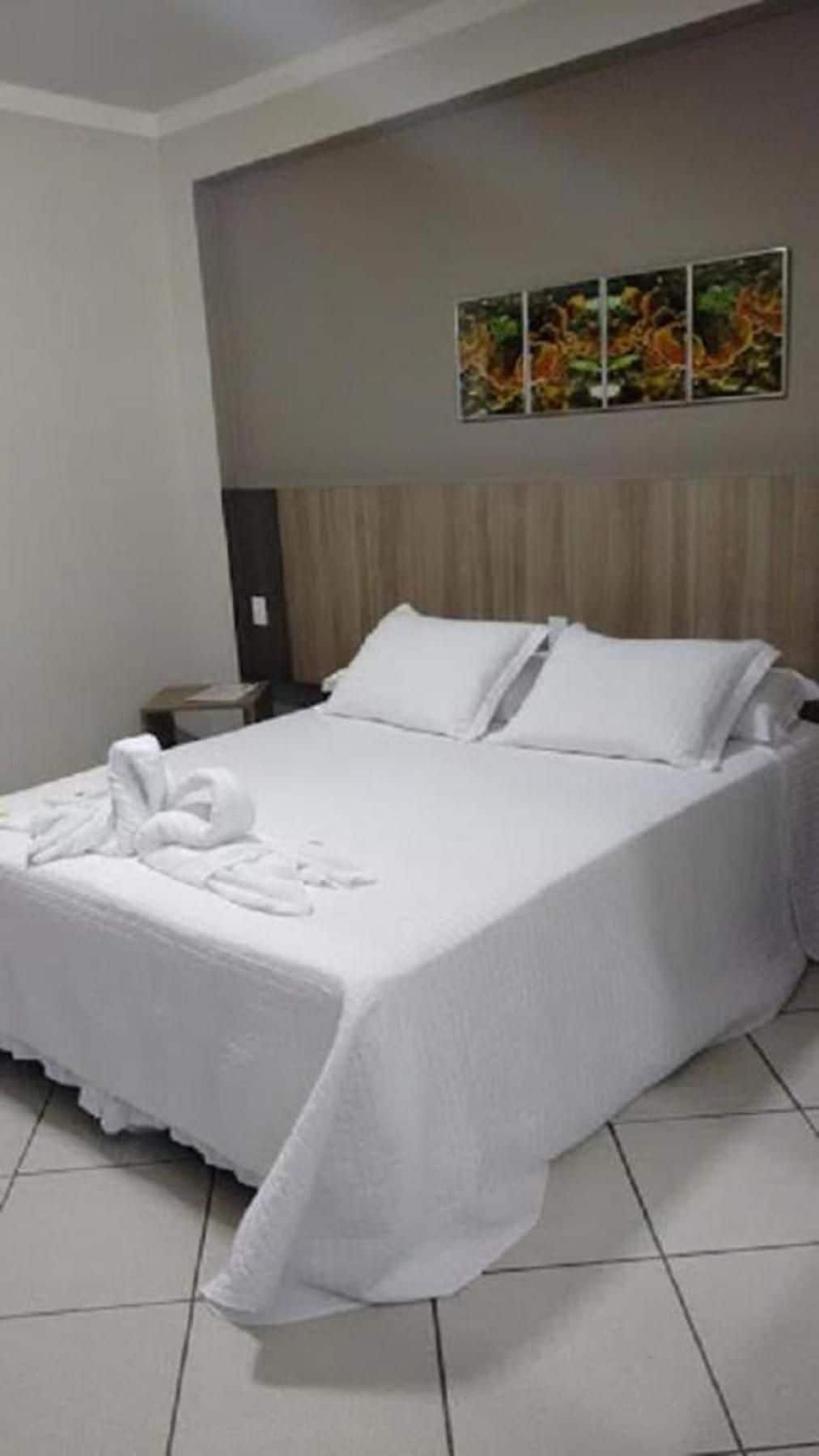 Quarto de hotel com cama bem arrumada, mesa de cabeceira, obra de arte na parede e chão de azulejos.