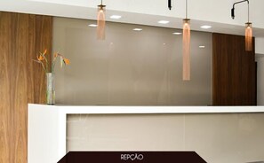 A reception desk with a modern design, featuring a wooden panel wall and a white counter. There are three pendant lights above the desk, and a vase with flowers on the left side.