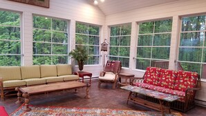 Una habitación acogedora con un banco de madera, un sofá rojo estampado y otro sofá verde estampado. Hay una mesita con un jarrón de flores y una jaula de pájaros colgando del techo.