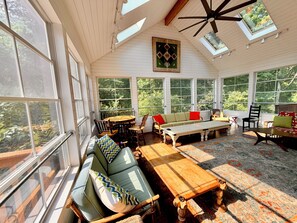 Une pièce baignée de lumière naturelle, avec un plafond en bois, de grandes fenêtres et un coin salon confortable comprenant un canapé et des fauteuils.