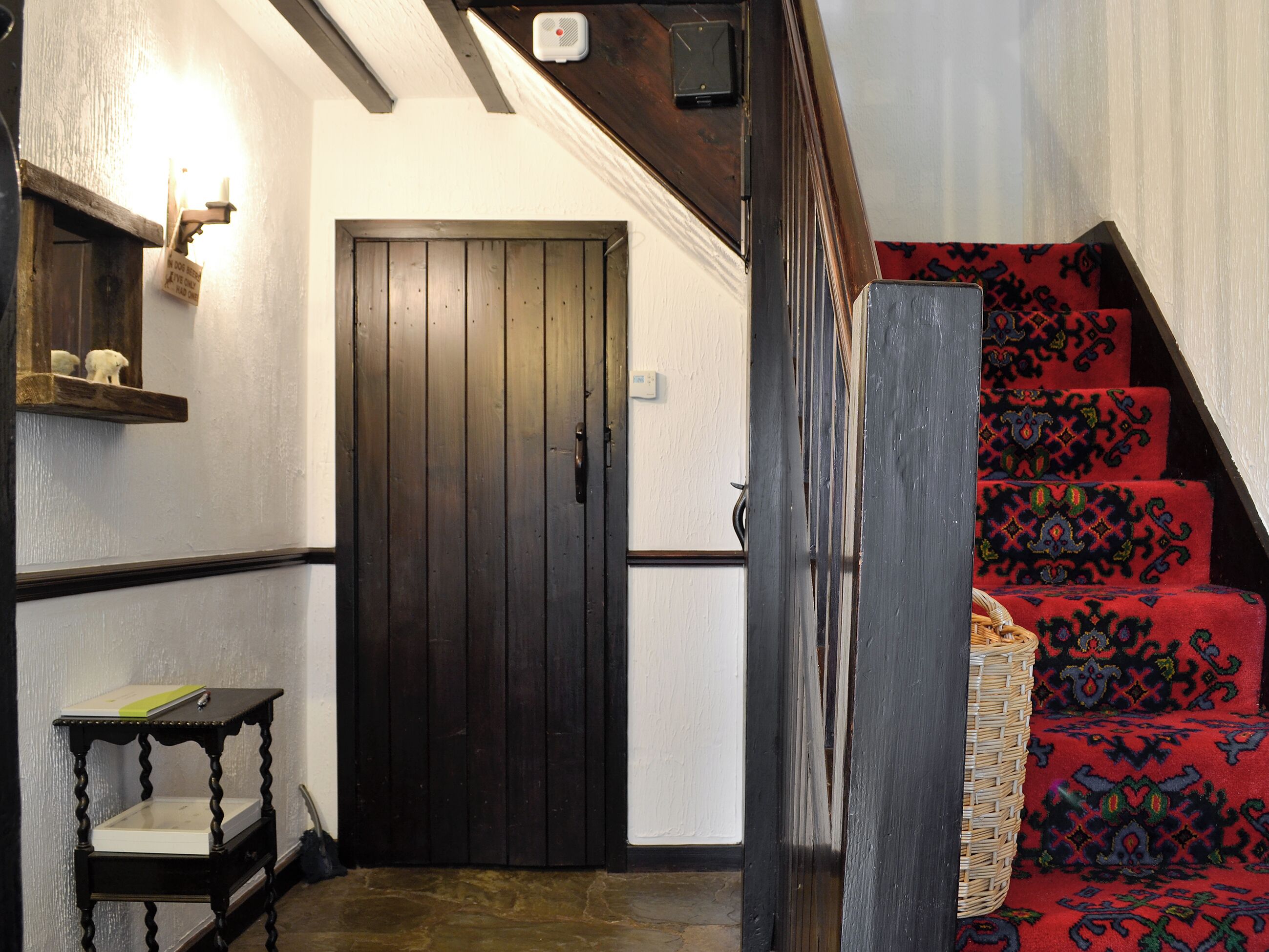 Una escalera con alfombra roja estampada, una puerta de madera oscura, una mesita con un libro y un estante fijado a la pared con una lámpara.