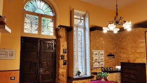 Una habitación con pared de piedra, puerta de madera, ventana con reja decorativa, candelabro y mesa cubierta con mantel rojo.