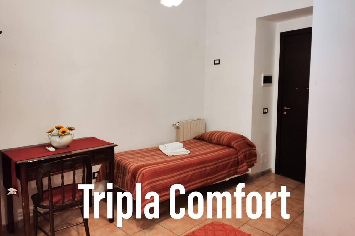 Una habitación pequeña y ordenada con una cama individual, una mesita con un florero y una silla.