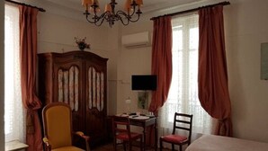 A room with a bed, two chairs, a desk with a computer, a chandelier, and curtains. - Hotel Cronstadt (Nice)