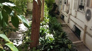 A narrow courtyard with a central tree, surrounded by various plants and a white building with shutters and a fan. - Hotel Cronstadt (Nice)