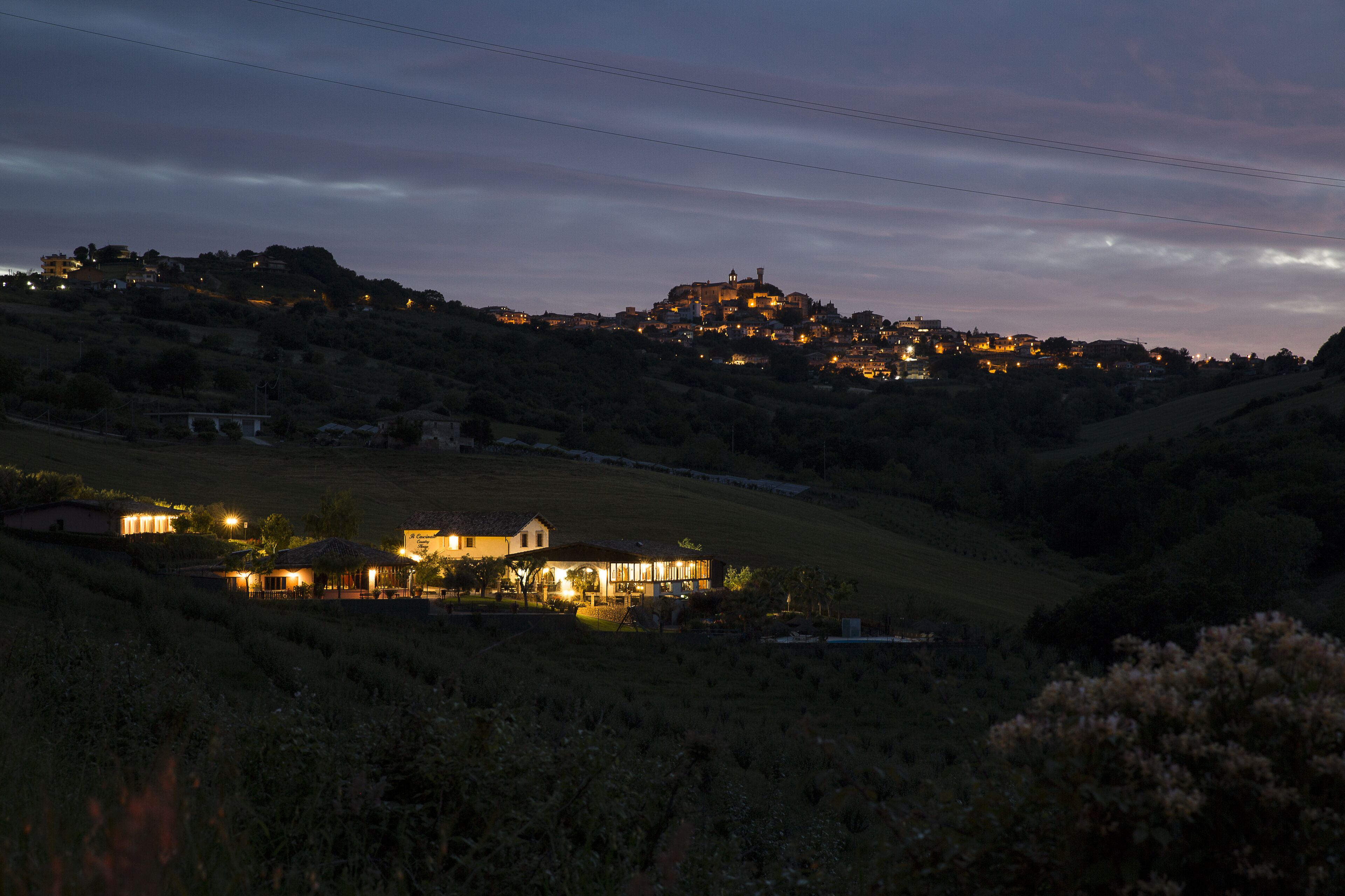 A rural landscape with a hilltop building and a lit house in the foreground.