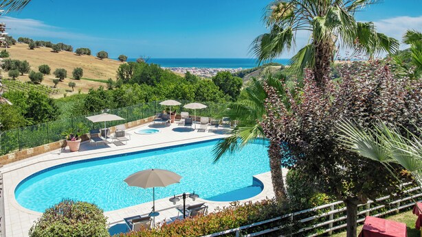 Piscina con sillones de descanso y sombrillas, rodeada de vegetación y con vistas a la costa.