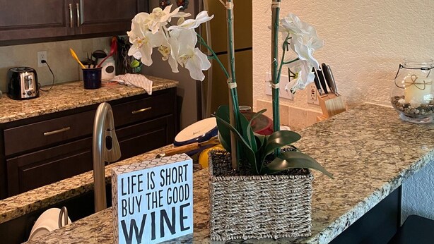 Eine Küche mit dunklen Holzschränken, einer Granitabdeckung und weißen Orchideen. Ein Schild zeigt die Aufschrift 'LIFE IS SHORT BUY THE GOOD WINE'.