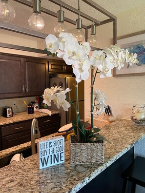 A kitchen with dark wooden cabinets, a granite countertop, and white orchids. There is a sign that reads "LIFE IS SHORT BUY THE GOOD WINE". - LONG TERM RENTALS...Sleeps 10...OFFERING DISCOUNTS…walk to Hotel & Amenities! (Horseshoe Bay)
