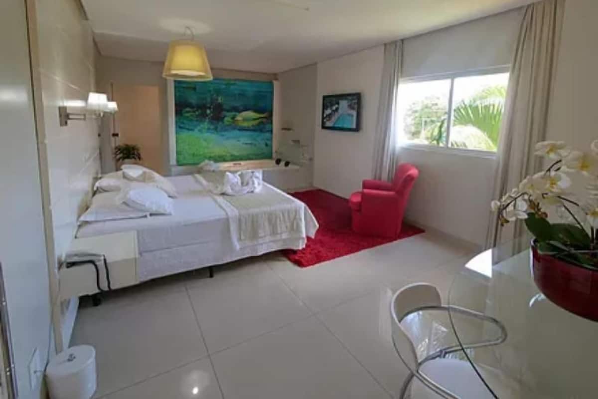 A hotel room with a bed, a desk with a chair, a red chair, a painting, and a window with curtains.