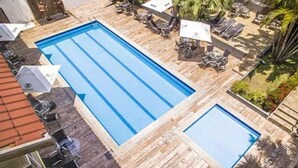 Una piscina rectangular con una terraza de madera alrededor, una piscina más pequeña y mobiliario exterior con sombrillas.