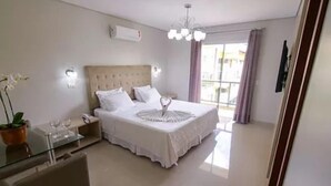 Habitación de hotel con una cama grande, una mesita de noche con lámpara, una pequeña mesa con dos copas de vino, una planta en maceta y vistas al exterior a través de un amplio ventanal.