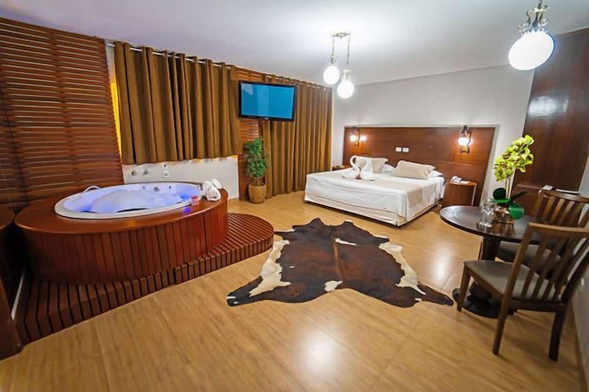 A modern hotel room with a large bed, a separate seating area with a table, a hot tub, and a flat-screen TV mounted on the wall.