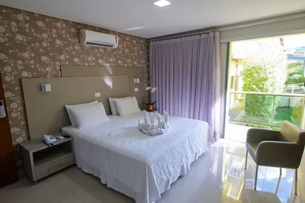 A hotel room with a large bed, a bedside table, an air conditioning unit, and a view of the outdoors.