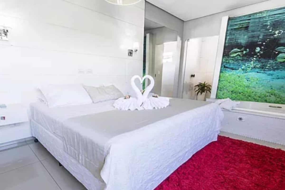 A modern bedroom with a large bed, white bedding, a red rug, and a wall-mounted aquarium.