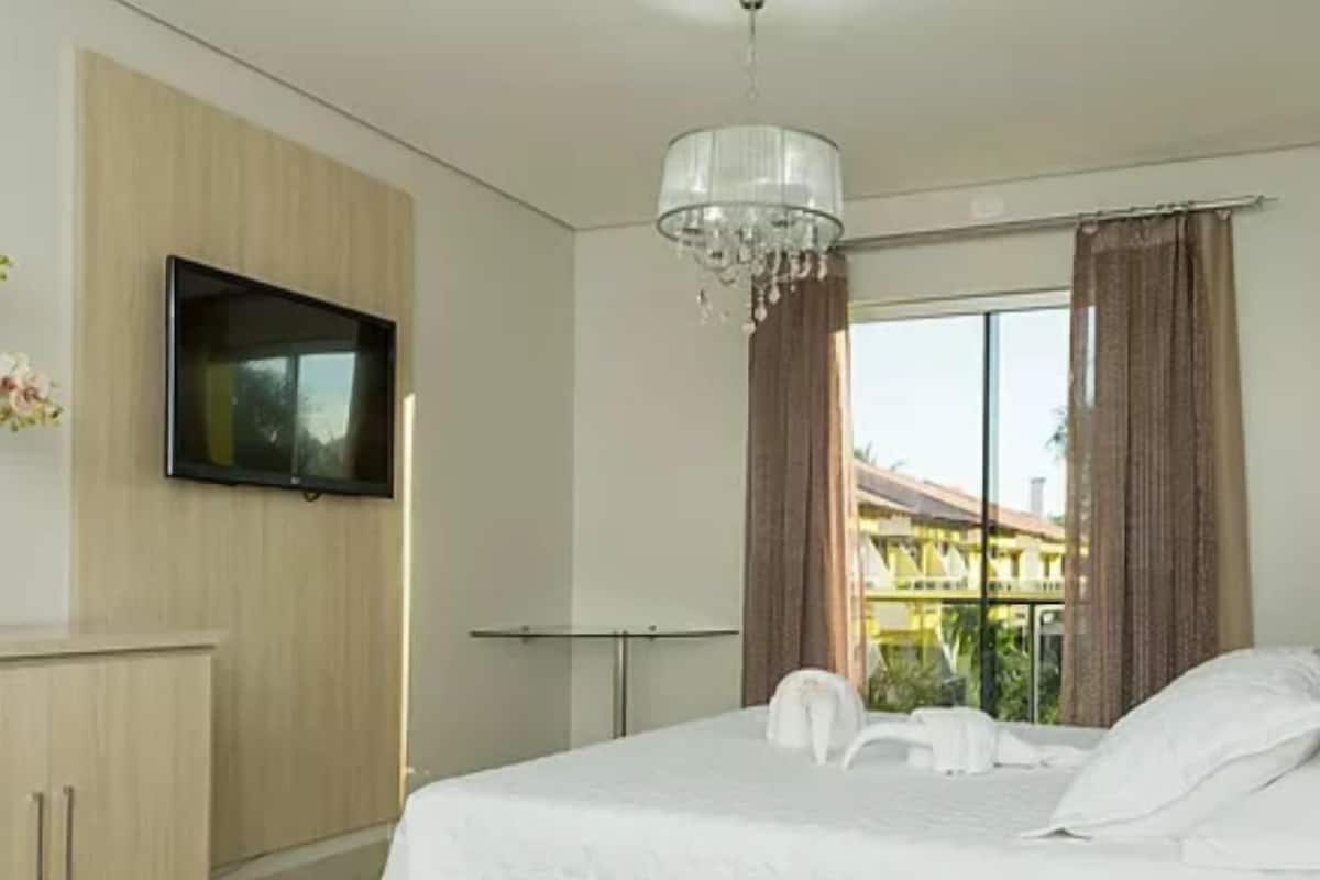 A hotel room with a bed, a TV mounted on the wall, a chandelier, and a window with curtains.