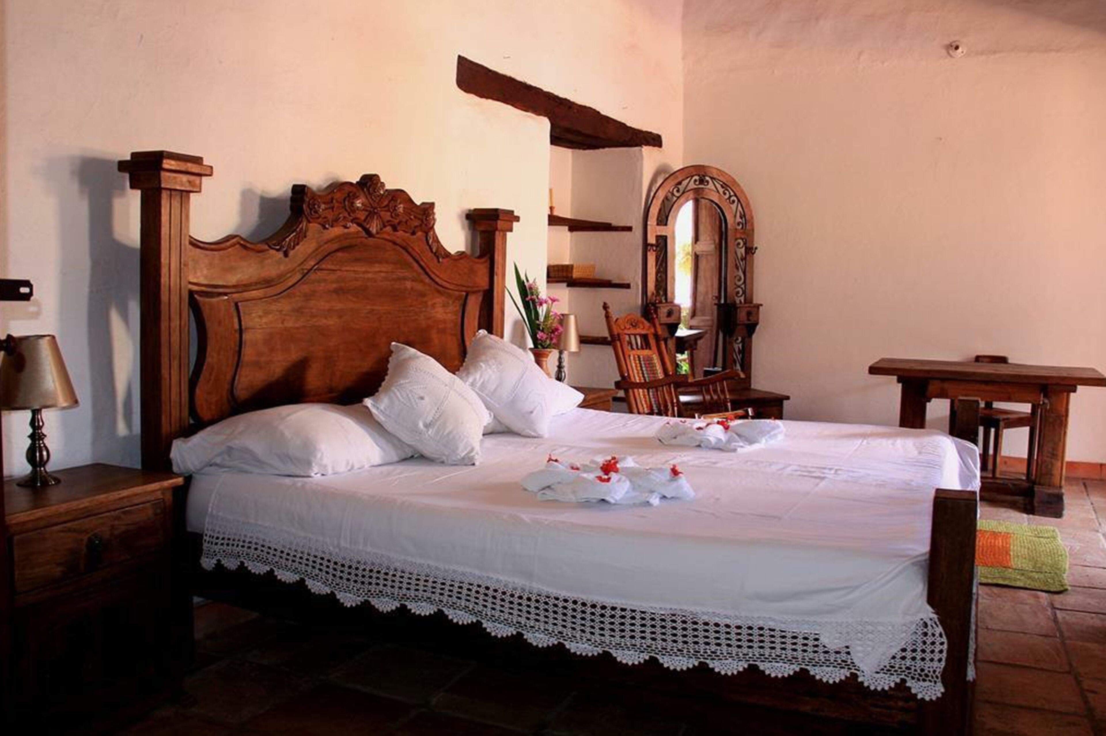 Una cama bien hecha con sábanas blancas y encaje, una mesita de noche de madera con una lámpara, una silla de madera, una pequeña mesa y un espejo en la pared.