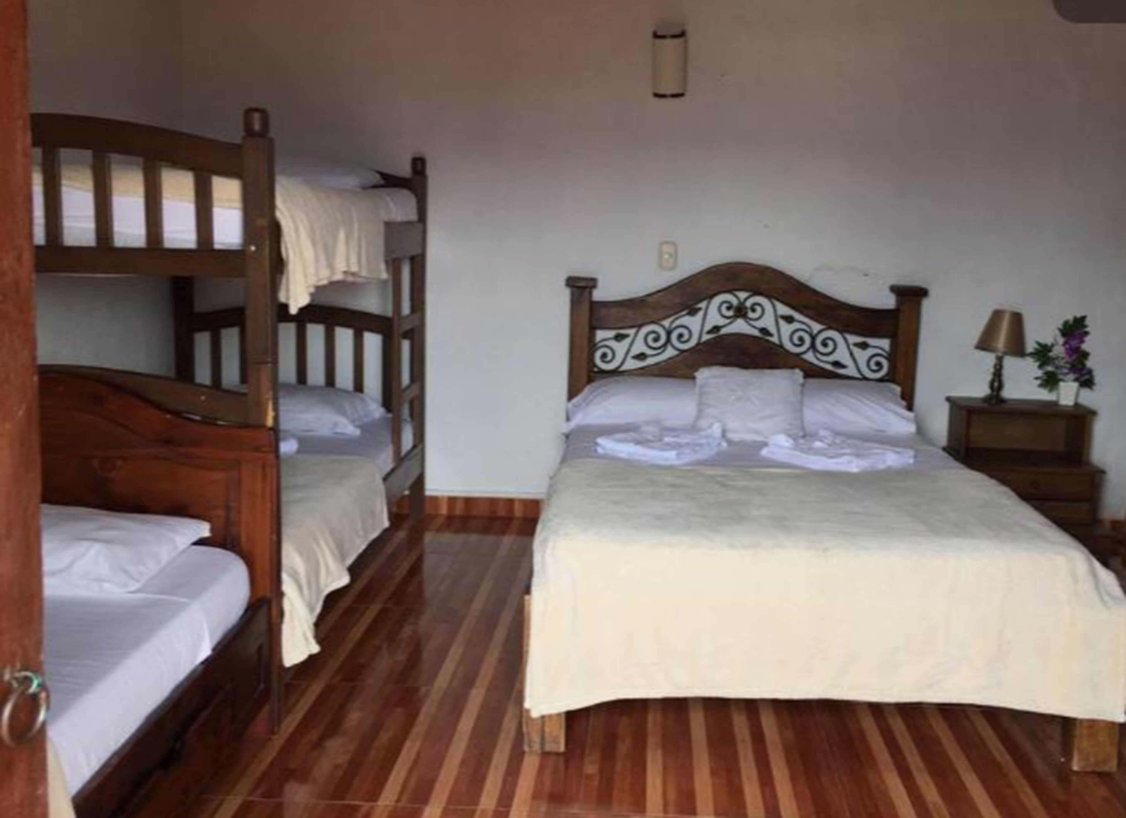 Una habitación con una cama grande, dos camas más pequeñas, suelo de madera, una mesita de noche con lámpara y una lámpara de pared.