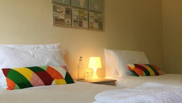 Habitación de hotel con dos camas, cada una con ropa de cama blanca y almohadas de colores. Hay una mesita de noche con una lámpara y un vaso de agua.