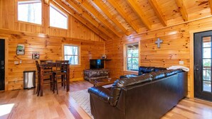 A log cabin interior with a dining area, a leather sofa, and a television.