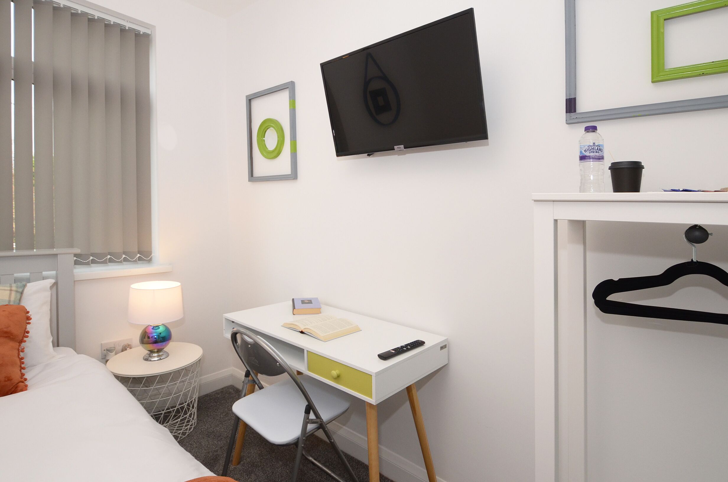 A bedroom with a bed, a desk with a chair, a TV, a window with blinds, and a water bottle on the nightstand.