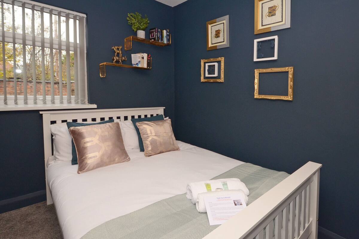 A neatly made bed with white and gold bedding, a white headboard, and a window with vertical blinds.