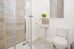 Une salle de bain avec une douche à parois de verre, un lavabo sur colonne, des toilettes et une petite étagère avec une plante.