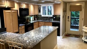 Une cuisine dotée de plans de travail en granit, de meubles en bois, d’une cuisinière noire et d’une fenêtre donnant sur des palmiers.