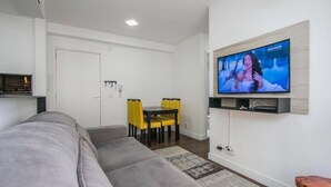 A modern living room with a grey sofa, a dining table with yellow chairs, and a mounted TV displaying a show. - Apartment next to the Grêmio Arena (Farrapos)